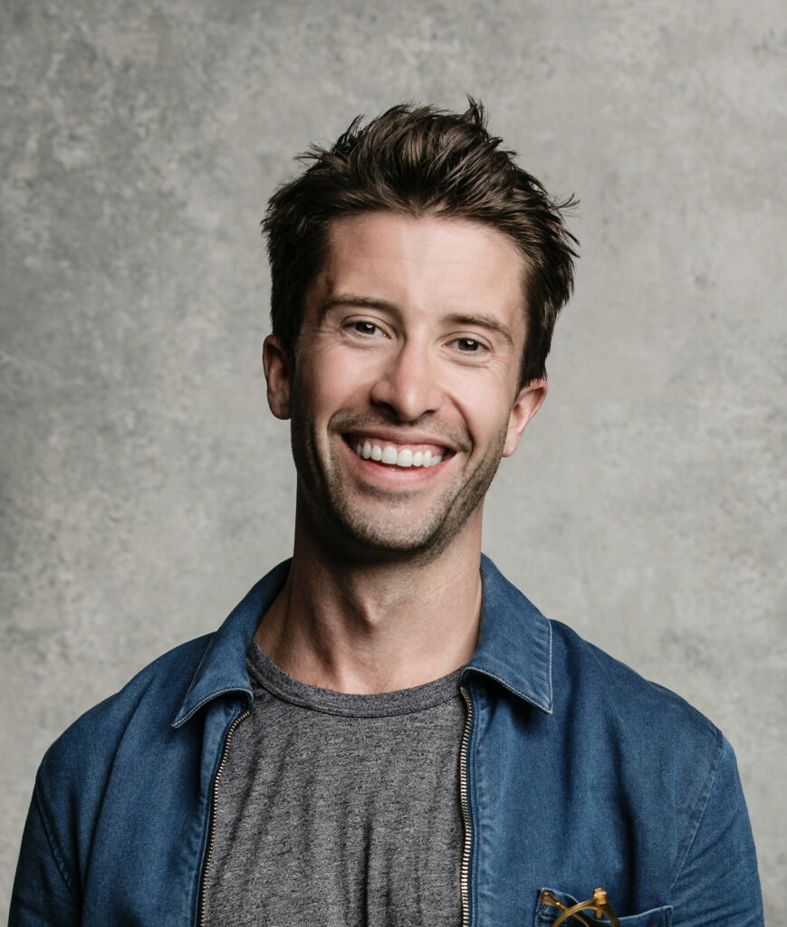 A man with short brown hair smiles, wearing a gray t-shirt and a blue denim button-up shirt against a gray background.