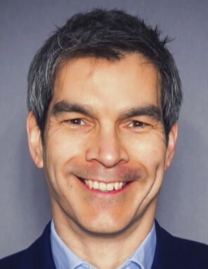 Smiling man with short gray hair wearing a blue shirt and dark jacket, in front of a gray background.