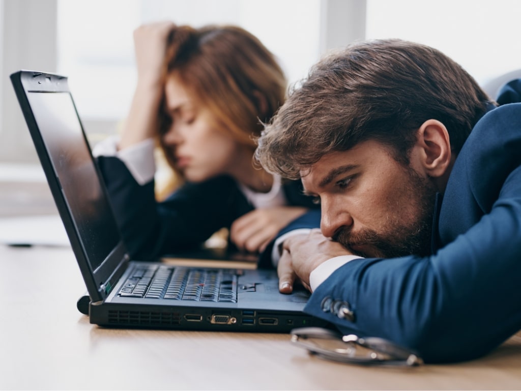 Two individuals in business attire rest their heads on a laptop, appearing frustrated or tired.