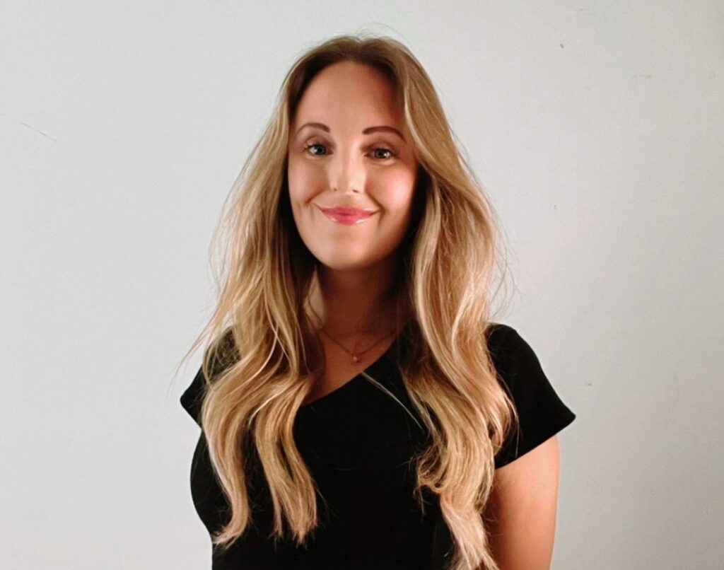 Woman with long hair stands against a plain background, wearing a black top and patterned skirt, smiling at the camera.