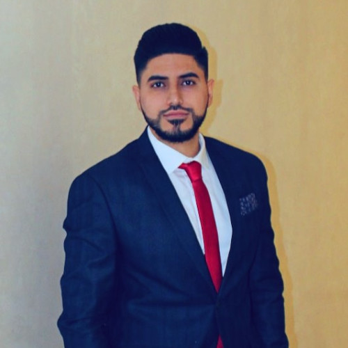 A man with short dark hair and a trimmed beard is wearing a dark suit, white shirt, and red tie, standing against a plain beige background.