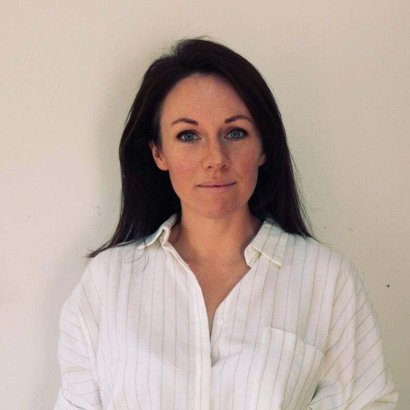 A woman with long brown hair wearing a white, collared, striped shirt stands in front of a plain light-colored background, looking directly at the camera.