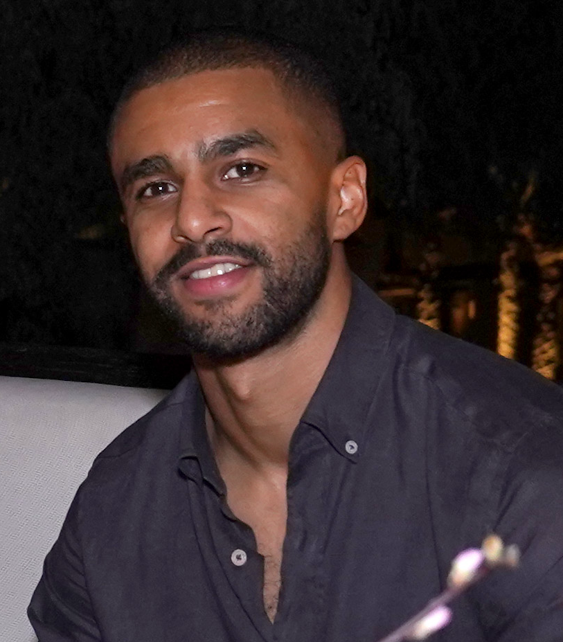 A man with short hair and a beard, wearing a dark button-up shirt, sits and smiles at the camera in a dimly lit setting.