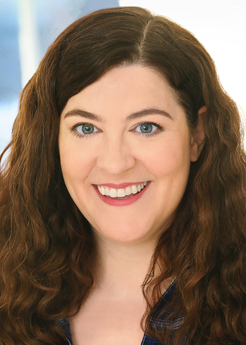 A woman with long, wavy brown hair and blue eyes smiles at the camera. She is wearing a blue top and is shown from the shoulders up against a light background.