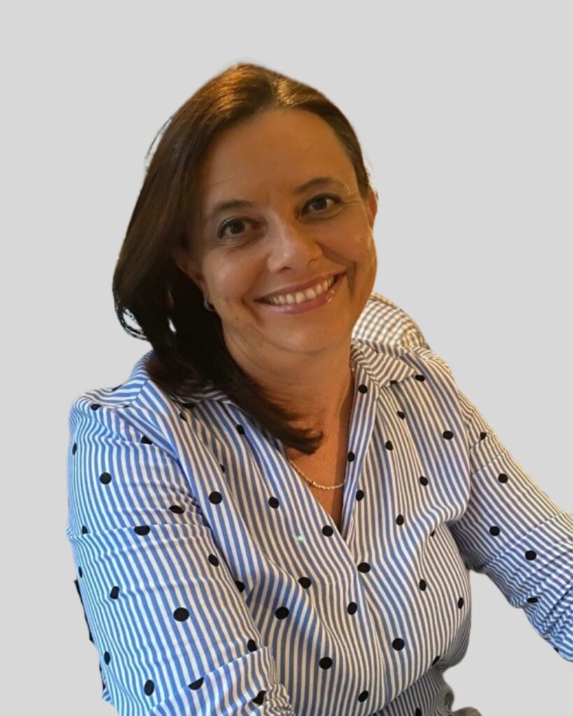 A woman with brown hair smiles while wearing a blue and white striped shirt with black polka dots, seated against a light grey background.