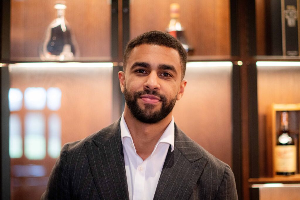 A man with short hair and a beard, wearing a pinstripe suit and white shirt, stands in front of a wooden shelf with bottles on display.